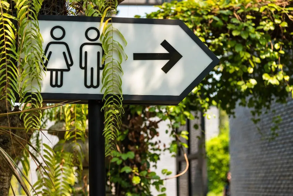 Panneau indiquant des toilettes hommes et femmes, flèche vers la droite dans un espace extérieur végétalisé